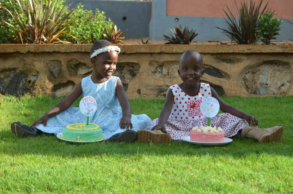 Two Maasai girls sitting on the grass in pretty floral dresses with their birthday cake placed in front of them.