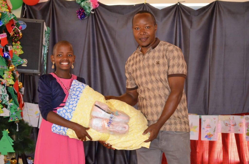 A smiling young Maasai girl and Elisante hold a christmas gift together in front of a beautifully decorated backdrop, celebrating the spirit of giving at MGRC